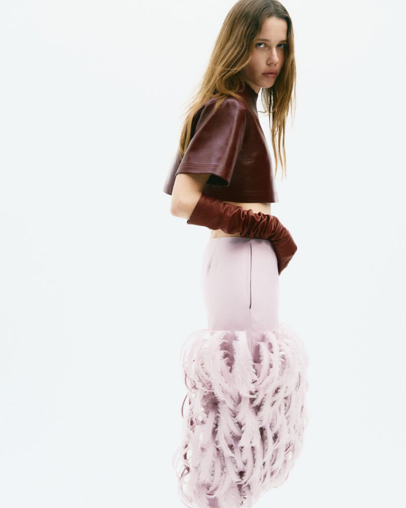 Model wearing a burgundy leather crop top, long gloves, and pink feathered skirt against a white studio background.