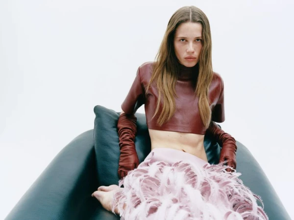 Model seated in a dark green armchair wearing a burgundy leather crop top, long leather gloves, and a pink feathered skirt.
