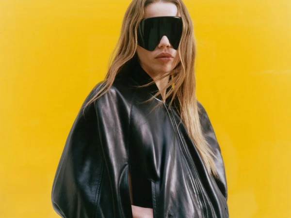 Model wearing a black leather jacket and large shield sunglasses against a vibrant yellow background