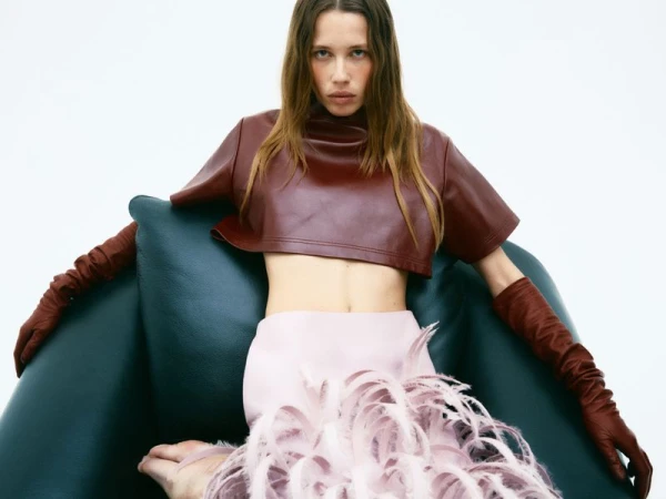Model seated in a green armchair wearing a burgundy leather crop top, pink skirt with feathered hem, and matching gloves against a white backdrop.