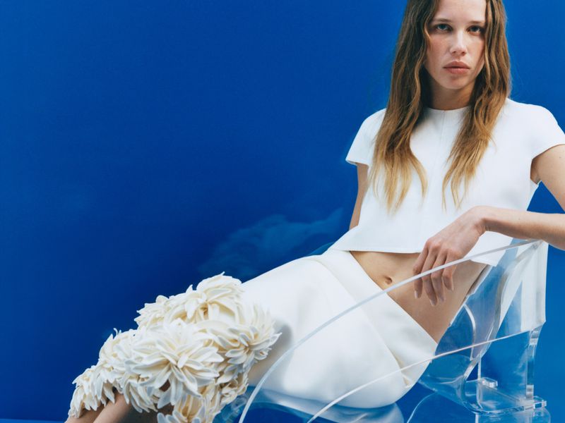 Model seated in a clear acrylic chair wearing a white top and textured floral skirt against a solid blue backdrop.