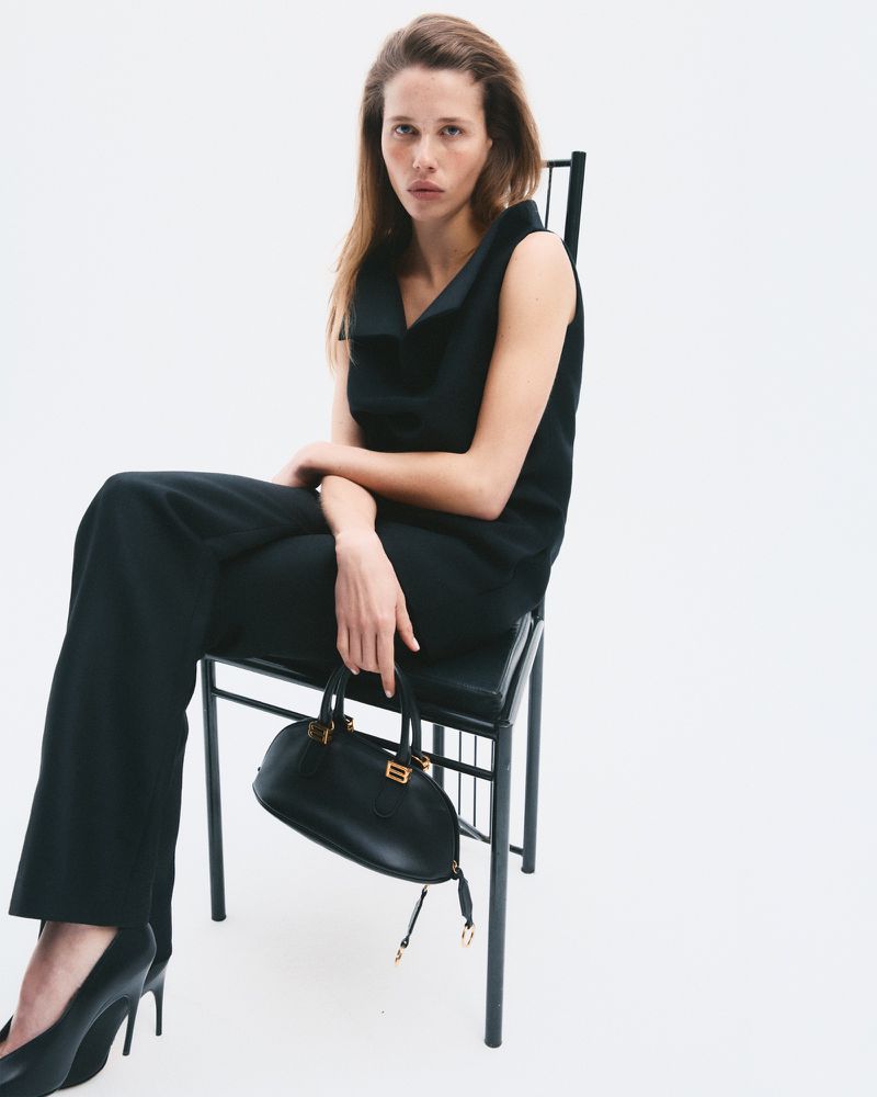 Model seated on a black chair wearing a black top and trousers while holding a small black handbag against a white background