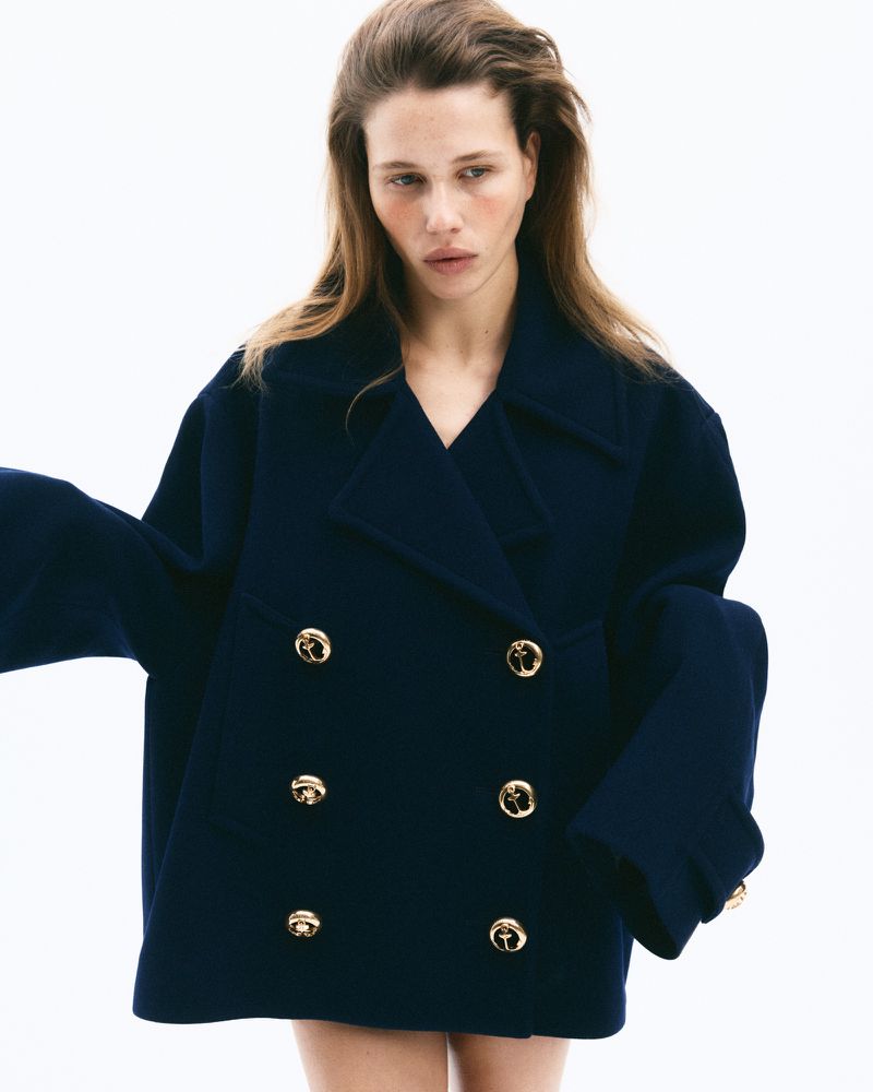 Model wearing a dark double-breasted coat with gold buttons against a white background for CR Fashion Book