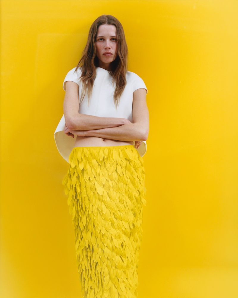 Model standing against a vibrant yellow background wearing a white top and a textured yellow feathered skirt.