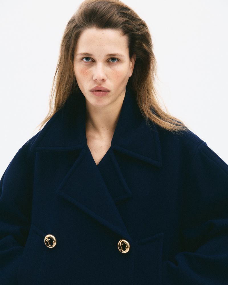 Model with long brown hair wearing a dark navy double-breasted coat against a plain white background for CR Fashion Book.
