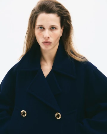 Model with long brown hair wearing a dark navy double-breasted coat against a plain white background for CR Fashion Book.