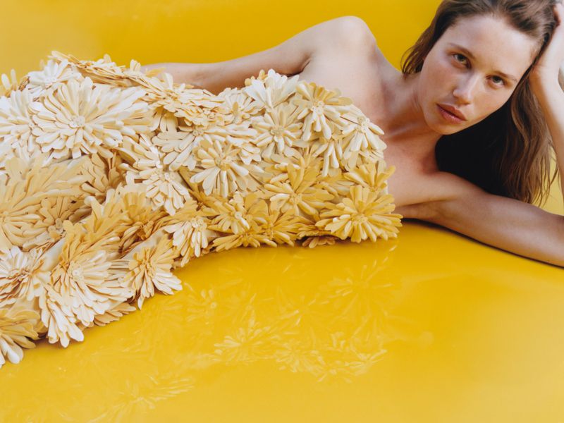 Model lying on a glossy yellow surface wearing a structured strapless dress covered in cream floral appliqués.