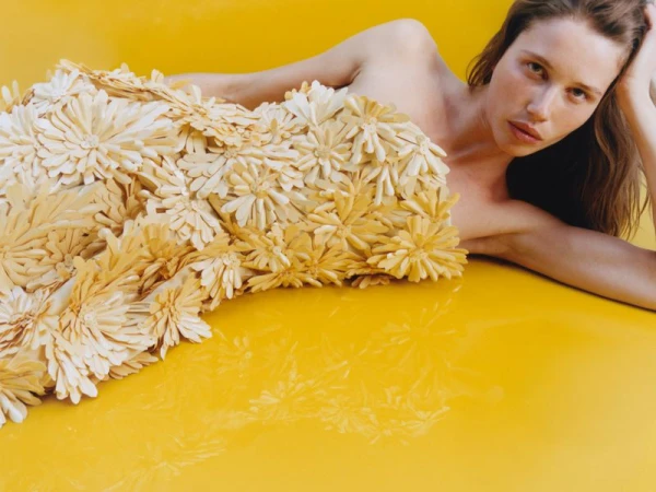 Model lying on a glossy yellow surface wearing a structured strapless dress covered in cream floral appliqués.
