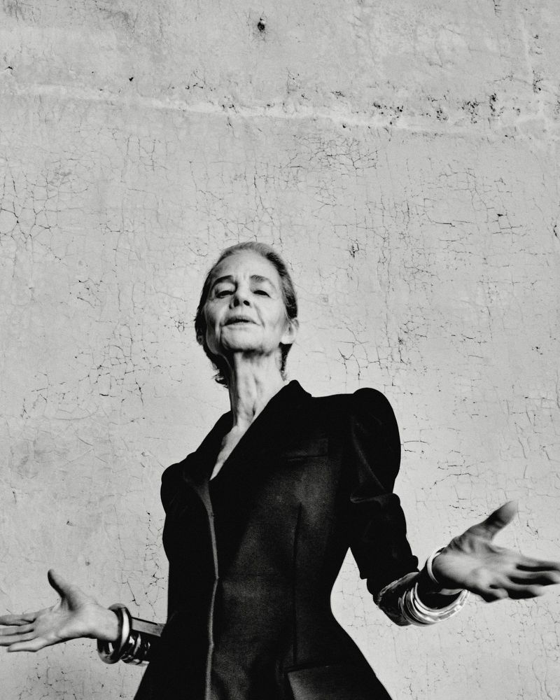Monochrome portrait of Charlotte Rampling wearing a dark structured jacket with wide gestures against a textured wall.