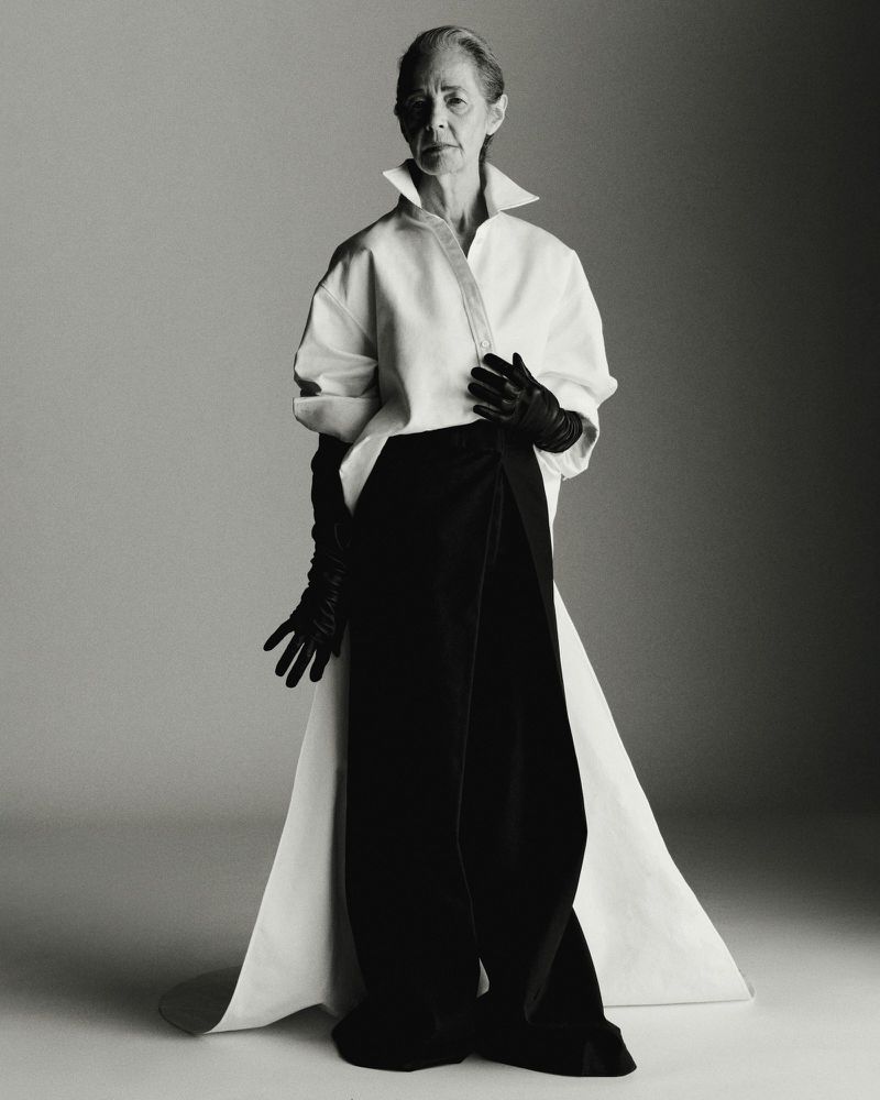 Elegant elderly woman poses in white top, black draped pants, and long black gloves against a neutral background in a studio fashion shot.
