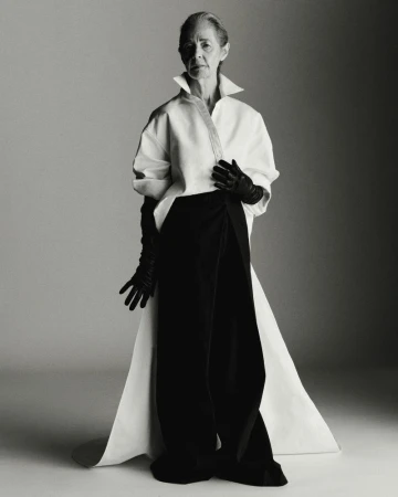 Elegant elderly woman poses in white top, black draped pants, and long black gloves against a neutral background in a studio fashion shot.