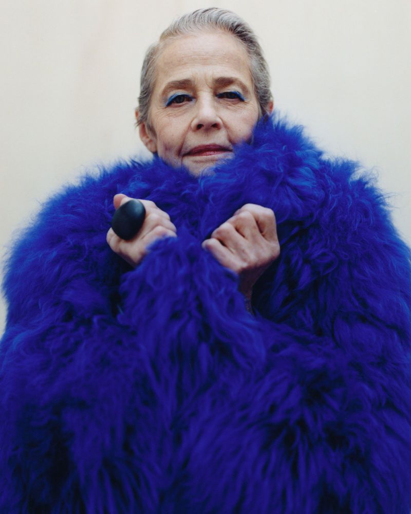 Elderly woman wearing a textured vibrant blue faux fur coat against a plain off-white background