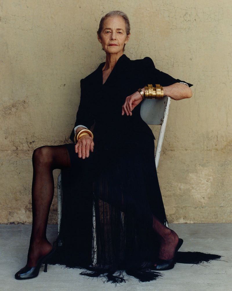 Elderly woman in a black jacket and skirt seated on a white chair against a weathered wall for an ICON Magazin Germany editorial