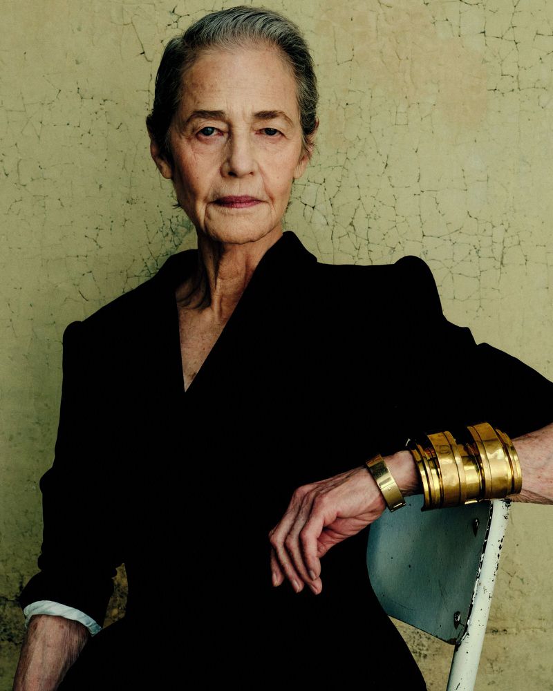 Portrait of Charlotte Rampling wearing a black top and gold cuffs seated against a textured green wall