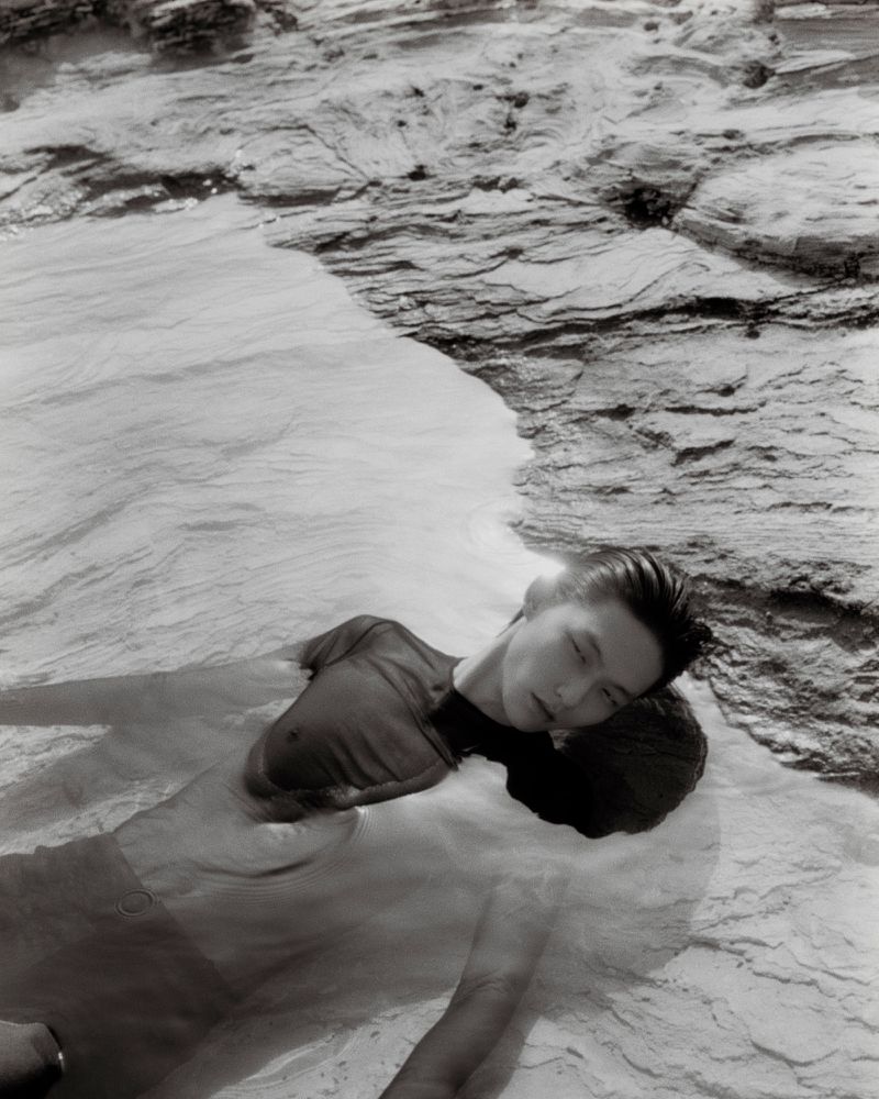Model floating in shallow water over a rocky seabed in a black and white editorial fashion photograph by Hordur Ingason.