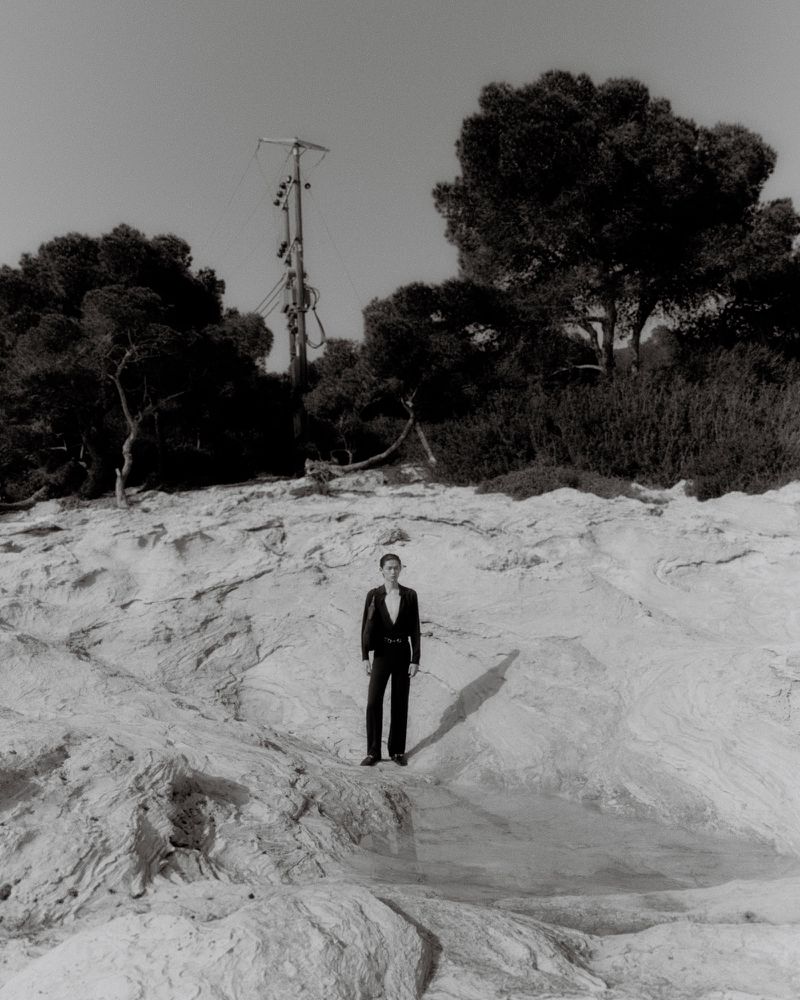 Model standing on rocky coastal terrain in a black suit against a backdrop of dark trees and a utility pole under natural light.