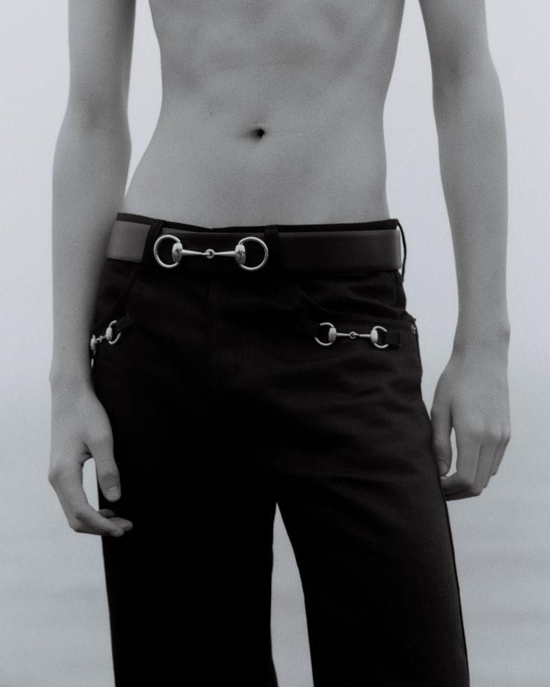 Close-up portrait of a model wearing dark trousers with a horsebit buckle belt against a pale grey background.