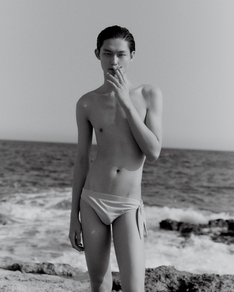 Black and white outdoor portrait of an Asian male model on a rocky beach wearing swimwear with ocean and sky in the background.