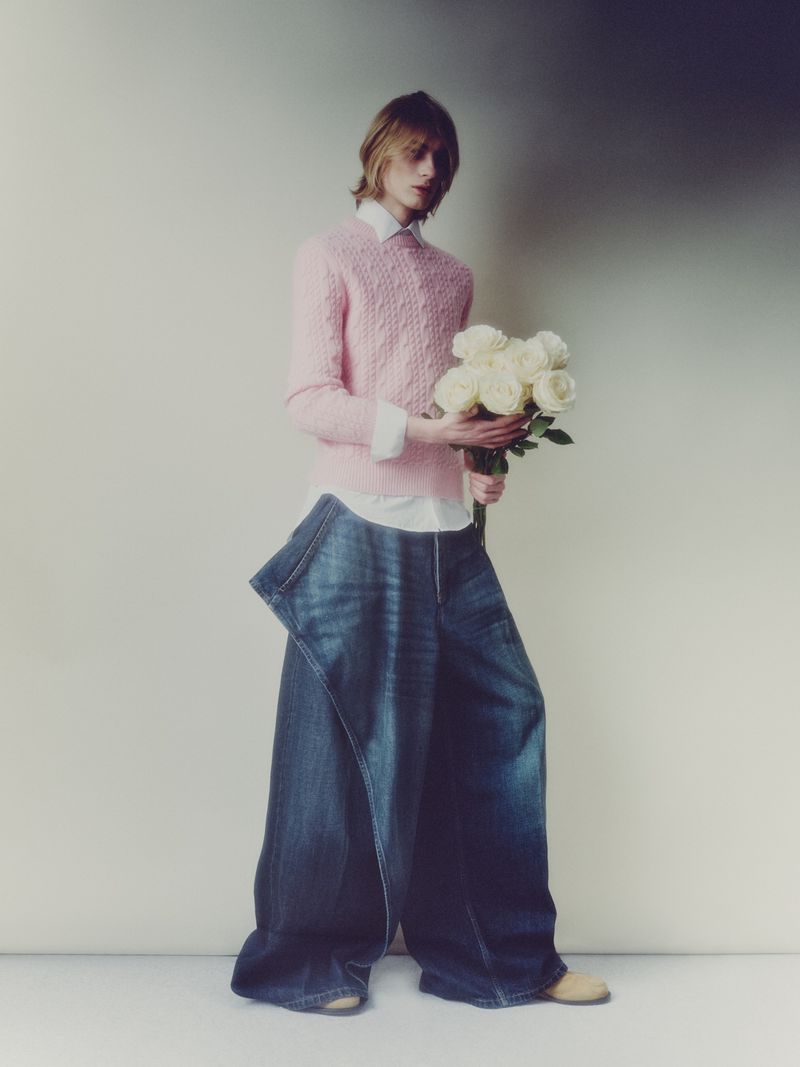 Model wearing a pink sweater and oversized denim pants holding a bouquet of white roses against a neutral studio backdrop.