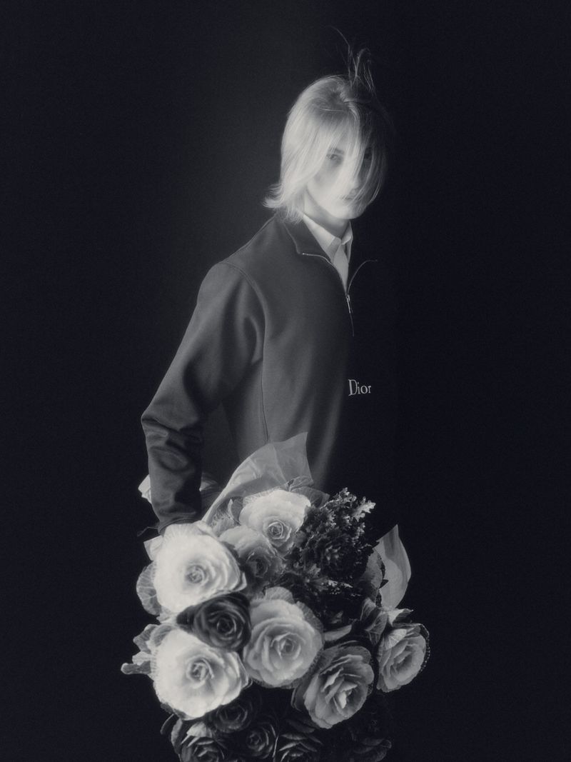 A person with blonde hair is holding a bouquet of roses in a studio with a black background. The model is wearing a jacket and a white collared shirt.