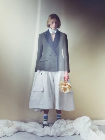 Model wearing a dark blazer and white flared skirt holding a floral bouquet against a soft gradient studio backdrop.