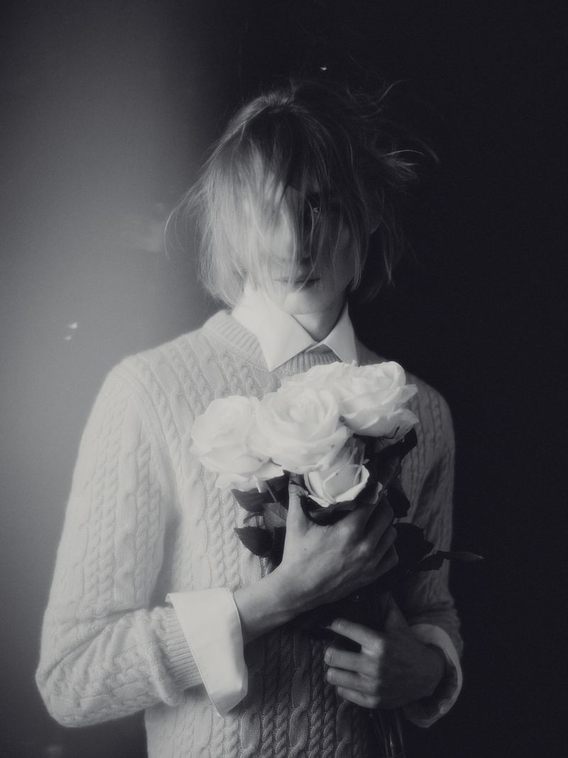 Model holding a bouquet of white roses wearing a knit sweater and collared shirt in a black and white editorial portrait