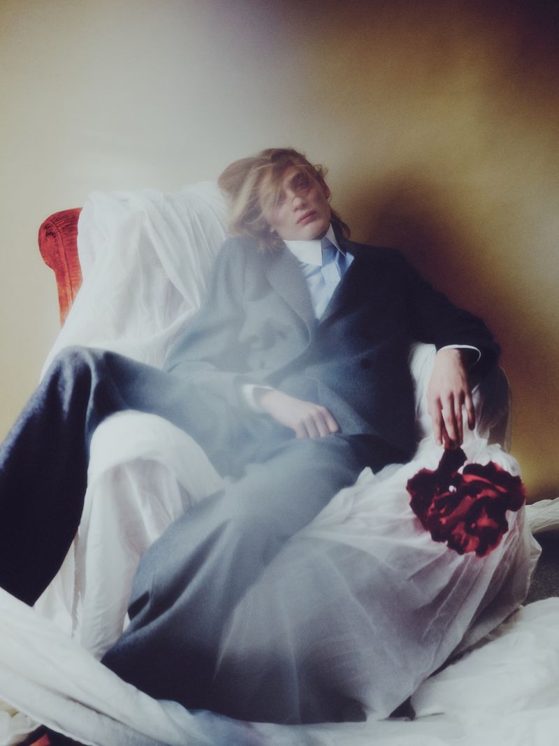 Model wearing a dark suit seated in an armchair draped with white fabric for a fashion editorial.