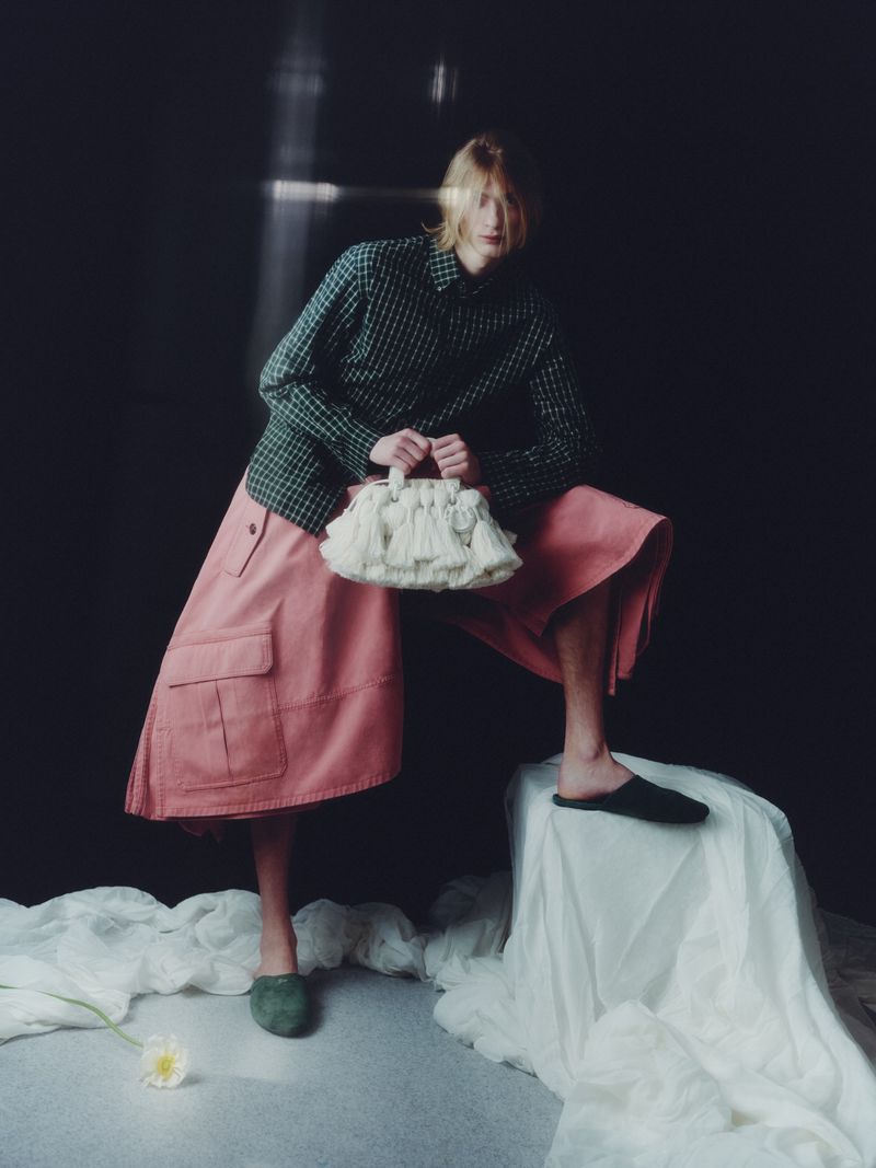 Model in plaid shirt and pink skirt holding a white fringed handbag against a dark backdrop