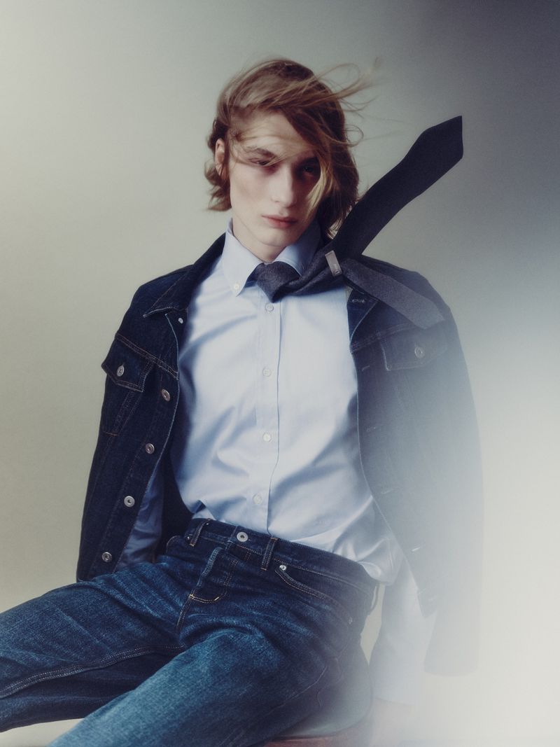 Model with long hair and flying tie wearing denim jacket and blue button-down shirt against plain studio background