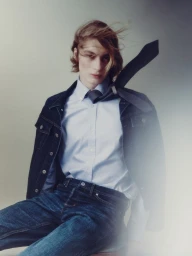 Model with long hair and flying tie wearing denim jacket and blue button-down shirt against plain studio background
