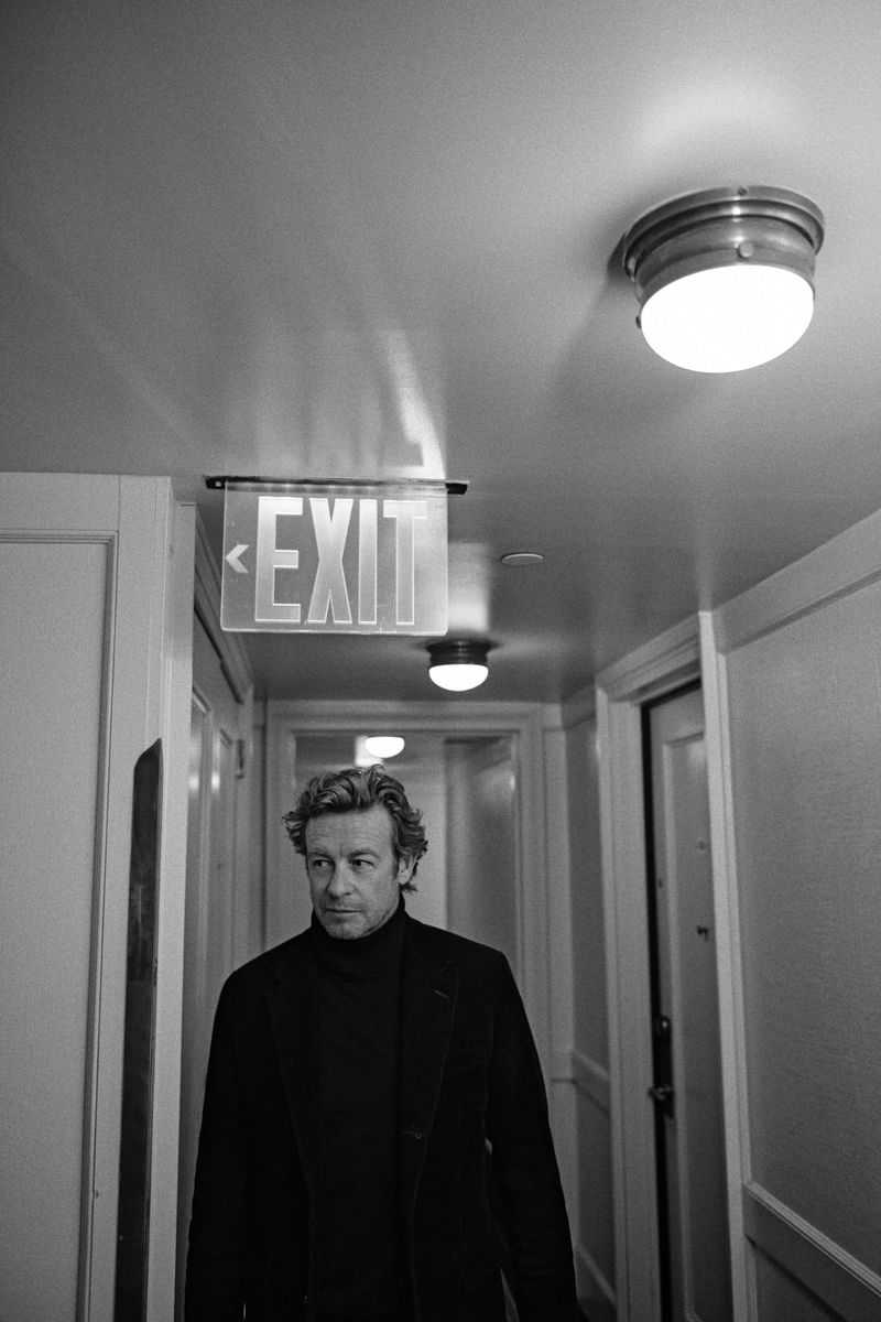 Indoor portrait of an adult male with dark hair wearing a black turtleneck and dark coat in a hallway with an exit sign.