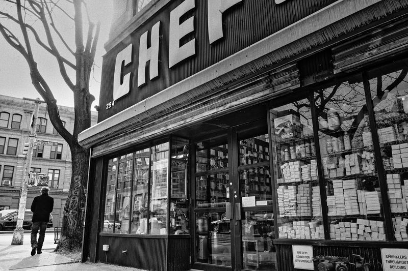Black and white photograph of a man walking past the storefront of CHEF Restaurant Supply store on a city street with bare trees.