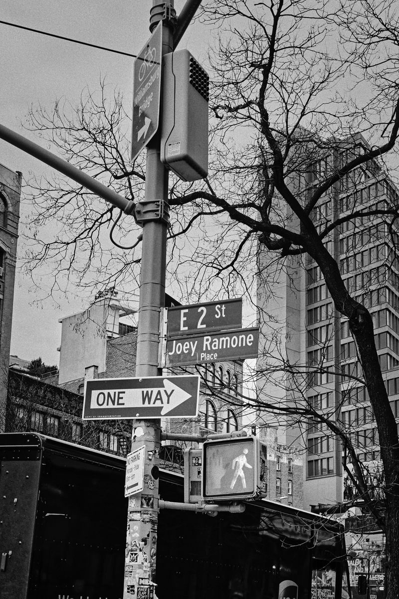 Black and white urban shot of a street corner with signs for E 2nd St, Joey Ramone Place, and a crosswalk signal showing a walking person.