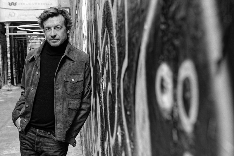 Black and white portrait of an adult male with brown hair wearing a dark turtleneck and suede jacket, leaning against graffiti-covered wall.