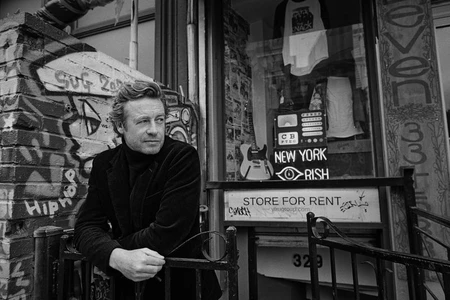 Black and white portrait of actor Simon Baker leaning against a graffiti-covered brick wall and metal railing in front of a storefront.