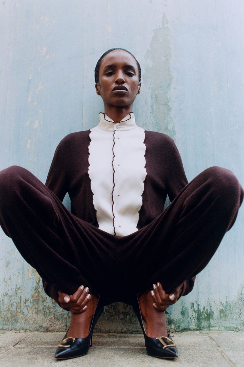 Model in a brown and white button-up top and trousers squatting against a weathered blue wall