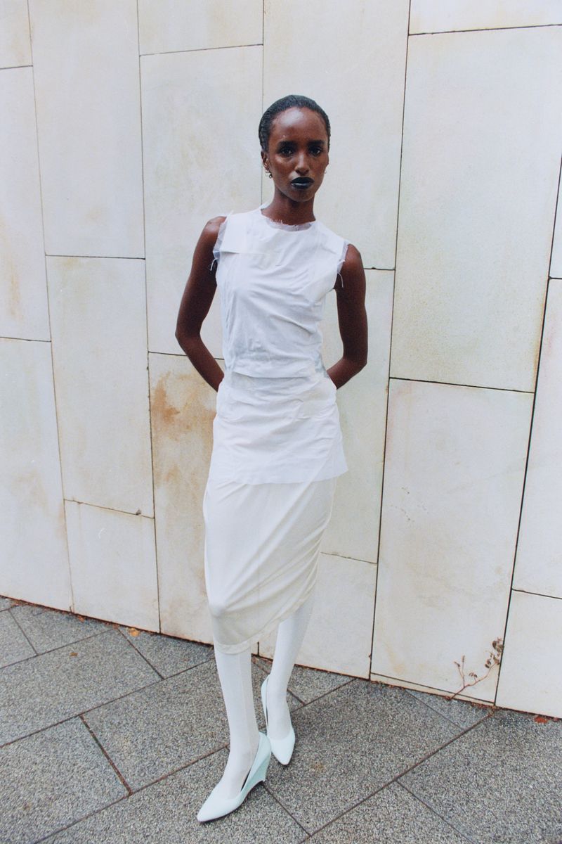 Model standing against a tiled stone wall wearing a white top, layered skirt, white tights, and white heels.