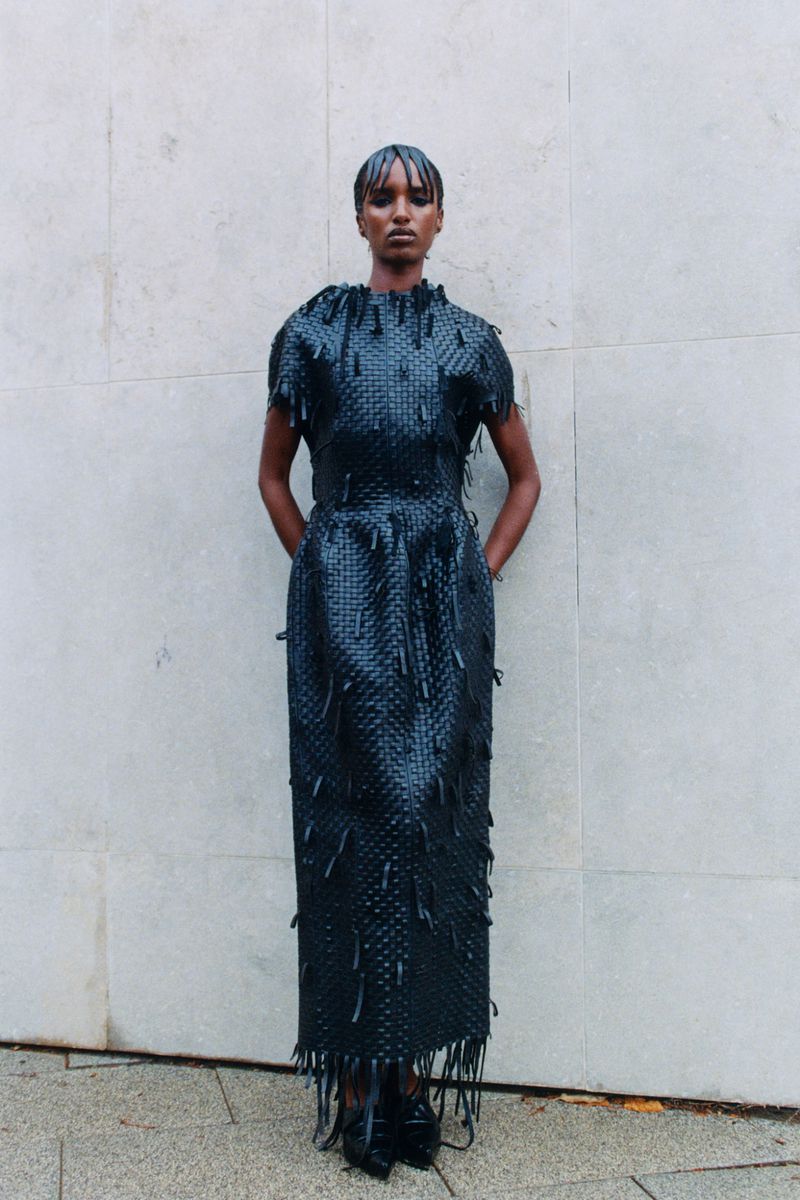Model wearing a dark textured dress with fringe details standing against a pale stone wall.