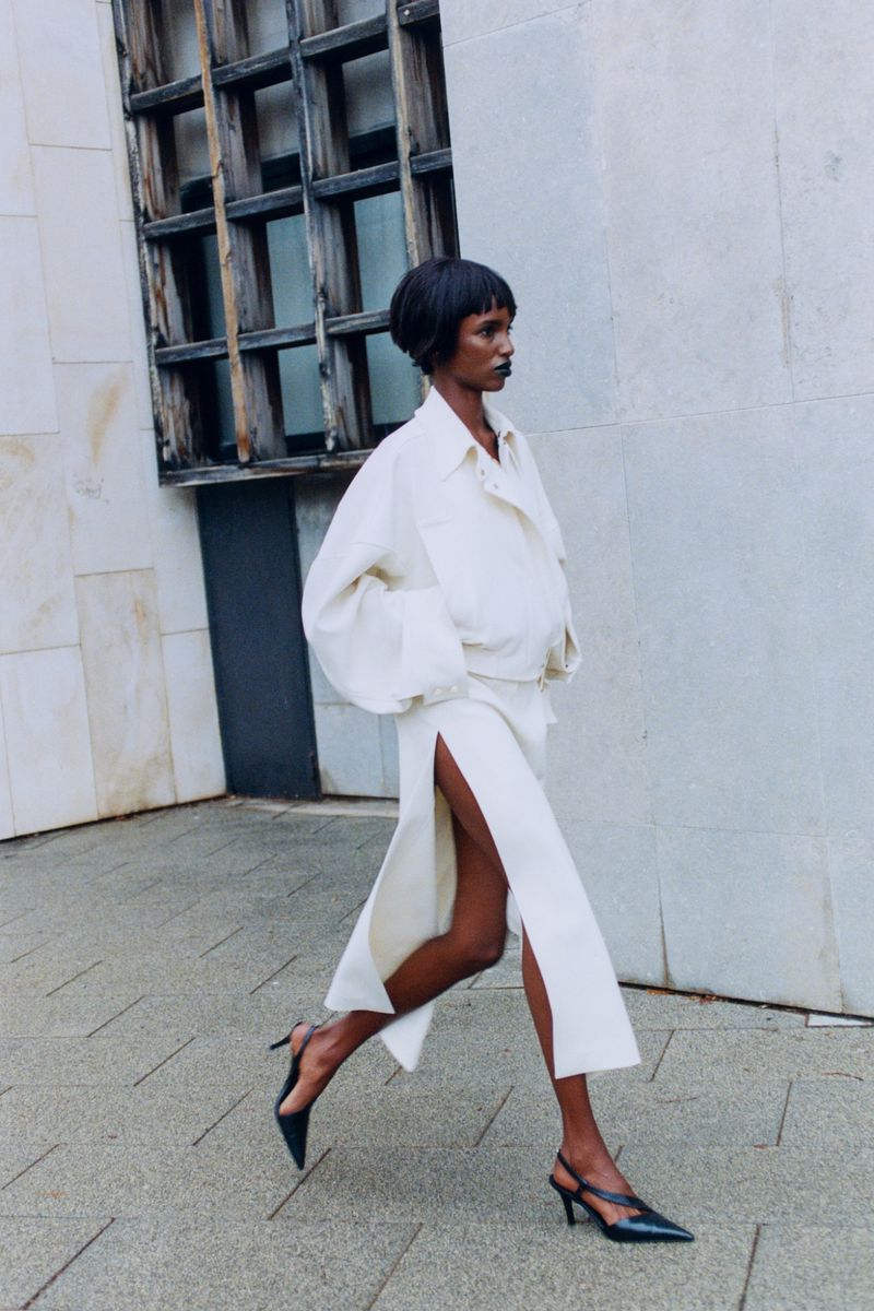A woman with dark skin and a short bob walks in a white jacket and skirt with black slingback heels against a light stone wall.
