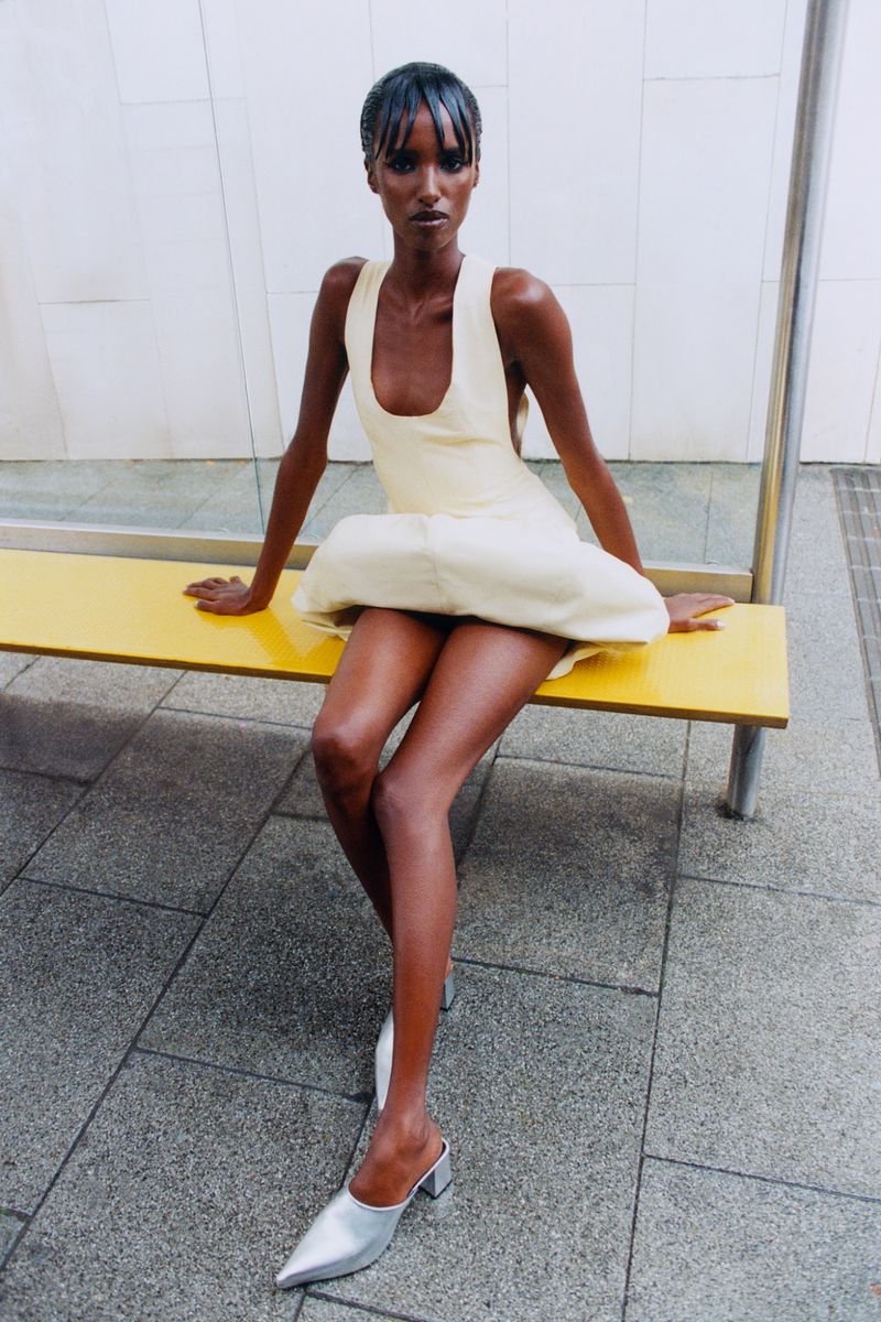 Model seated on a yellow bench wearing a cream scoop-neck dress and silver pointed-toe heels against a tiled wall