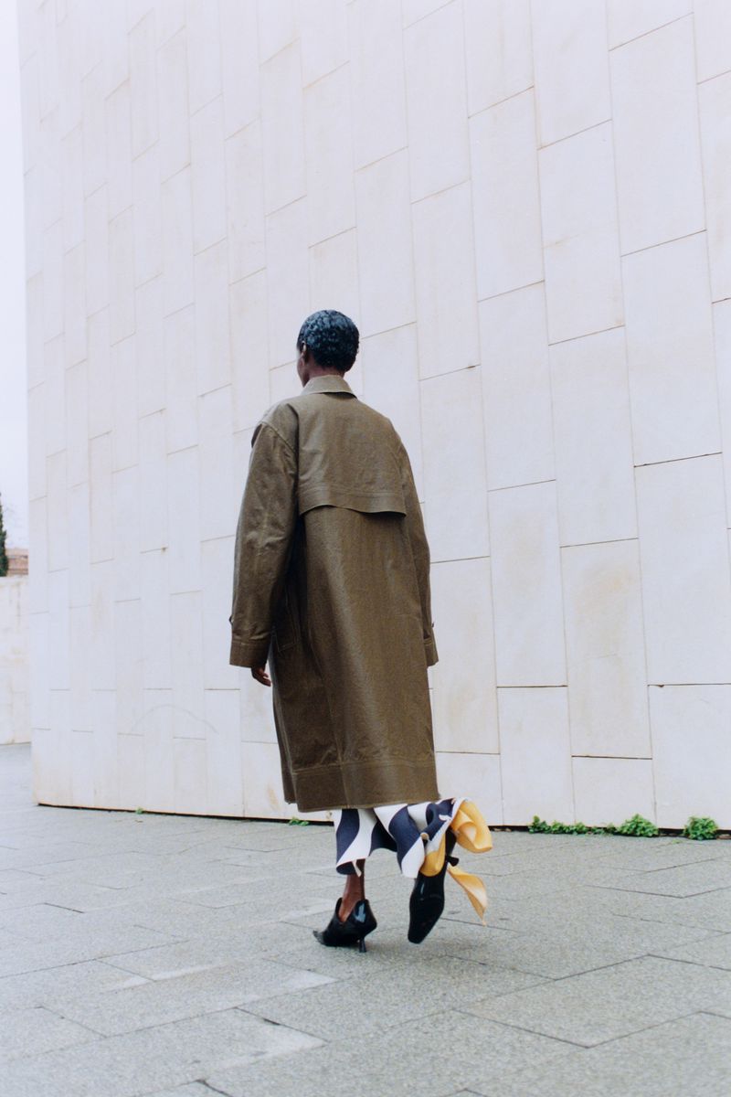 Fashionable person in a trench coat and black shoes walks in an outdoor setting with a white wall in the background.