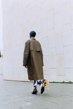 Fashionable person in a trench coat and black shoes walks in an outdoor setting with a white wall in the background.