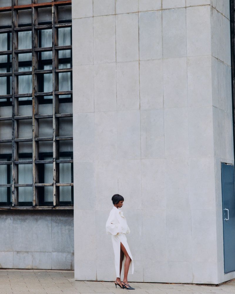 Model standing against a large concrete building exterior next to a gridded window facade for Manifesto Magazine
