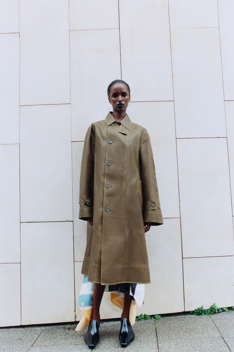 Model standing outdoors wearing an olive trench coat against a paneled stone wall