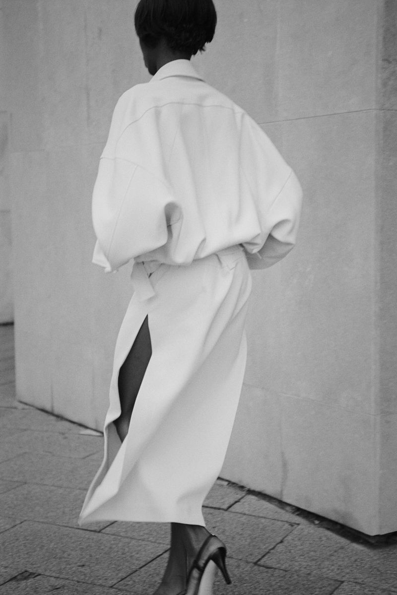 Model seen from behind wearing an oversized white button-down shirt and matching skirt with a side slit on an urban stone pavement.