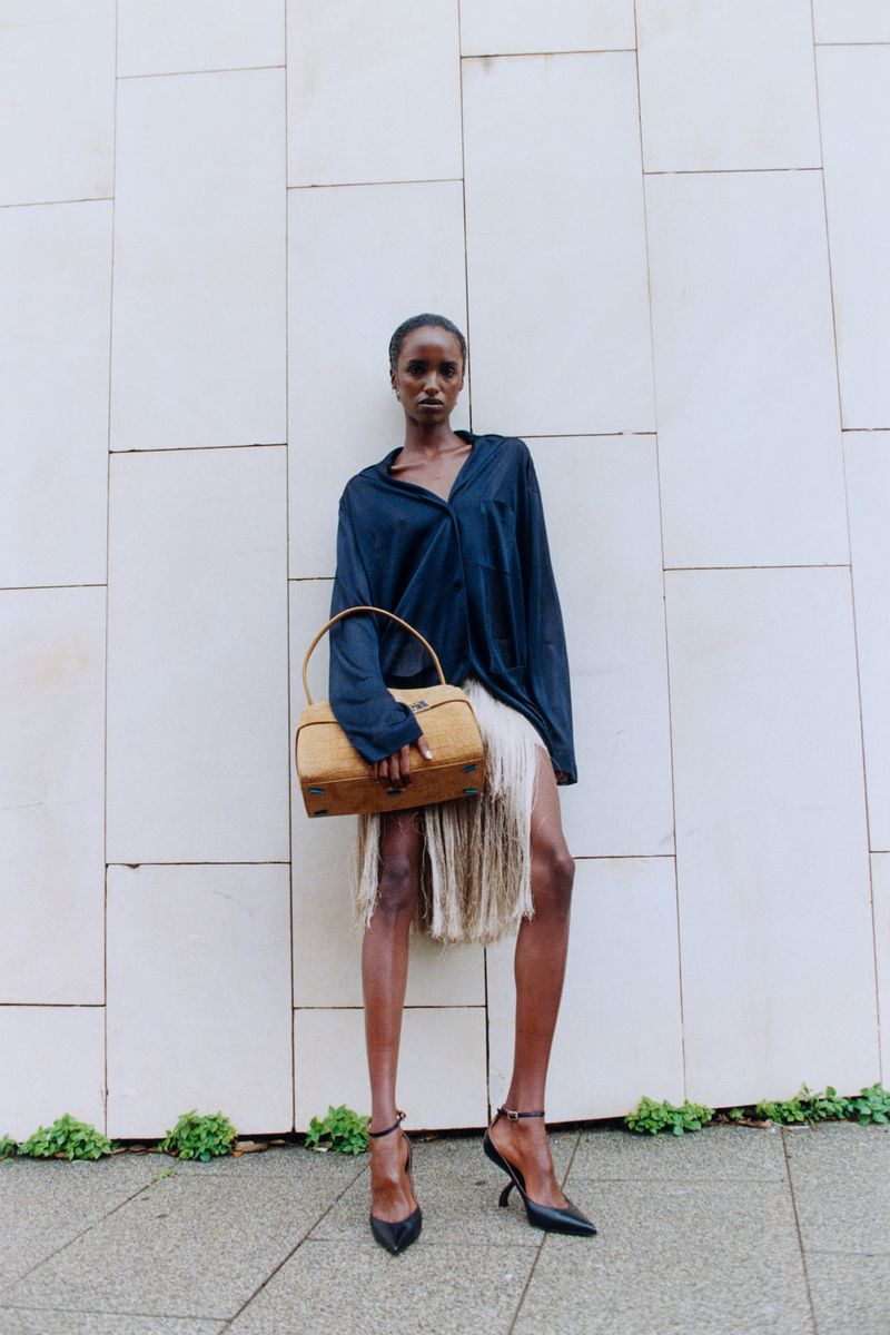 Model wearing a dark shirt and fringed skirt holding a tan handbag against a light tiled wall.