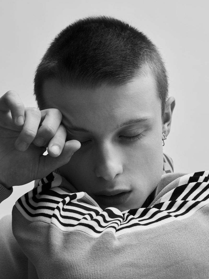 Close-up portrait of a young man with short hair wearing a striped sweater, with his hand near his face and eyes closed.