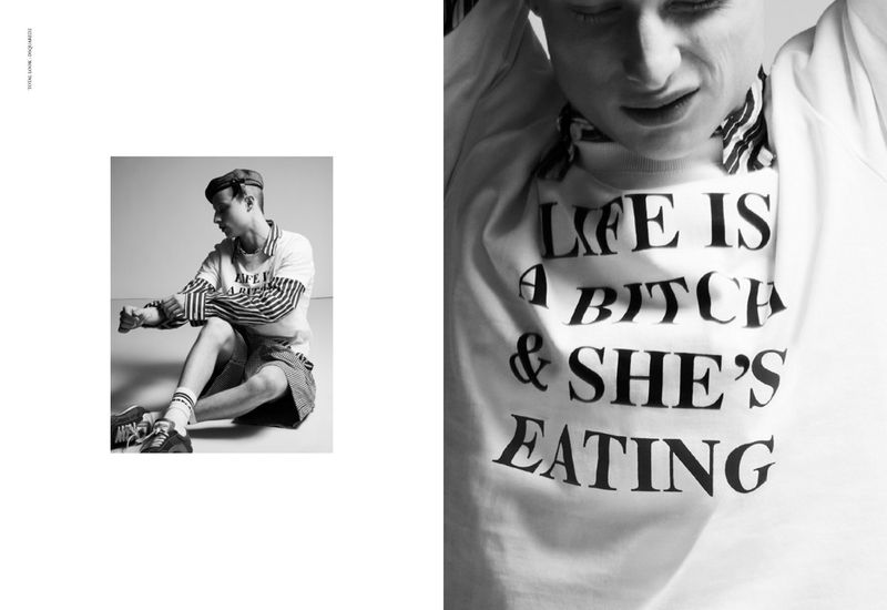Black and white editorial portrait of an actor wearing a graphic sweatshirt and cap seated against a seamless white studio background.