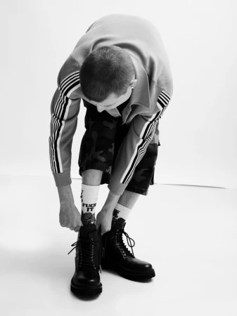 Monochrome portrait of a man leaning down to lace up his black combat boots against a plain white studio backdrop.