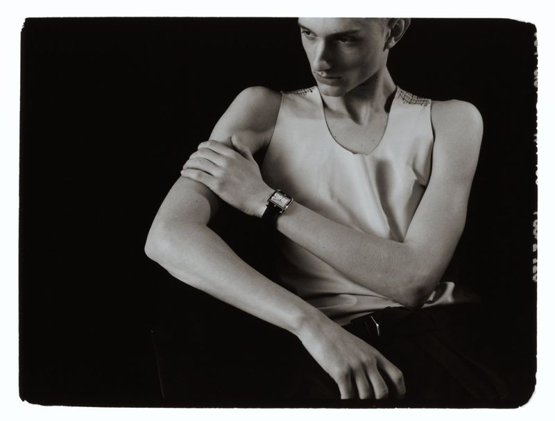 Model wearing a watch on a black strap and a light tank top against a dark background in an editorial portrait